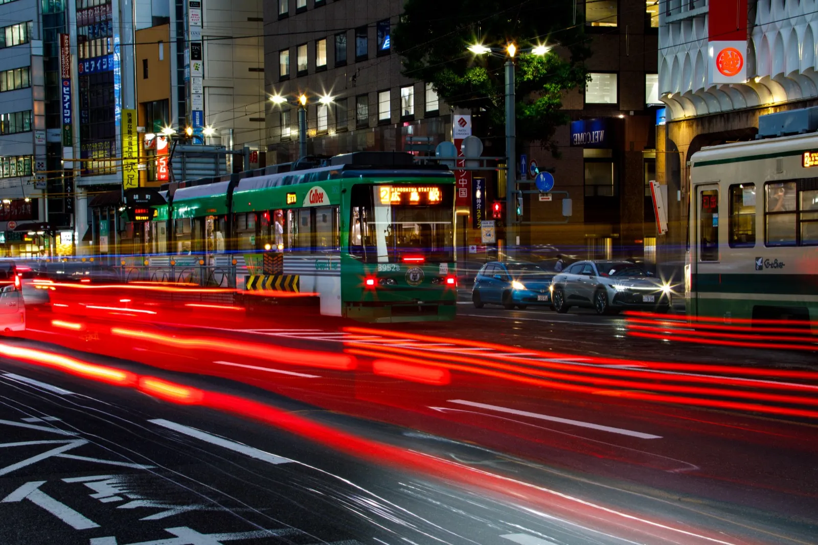 A green streetcar crossing a Hiroshima intersection at night with red light trails in the foreground