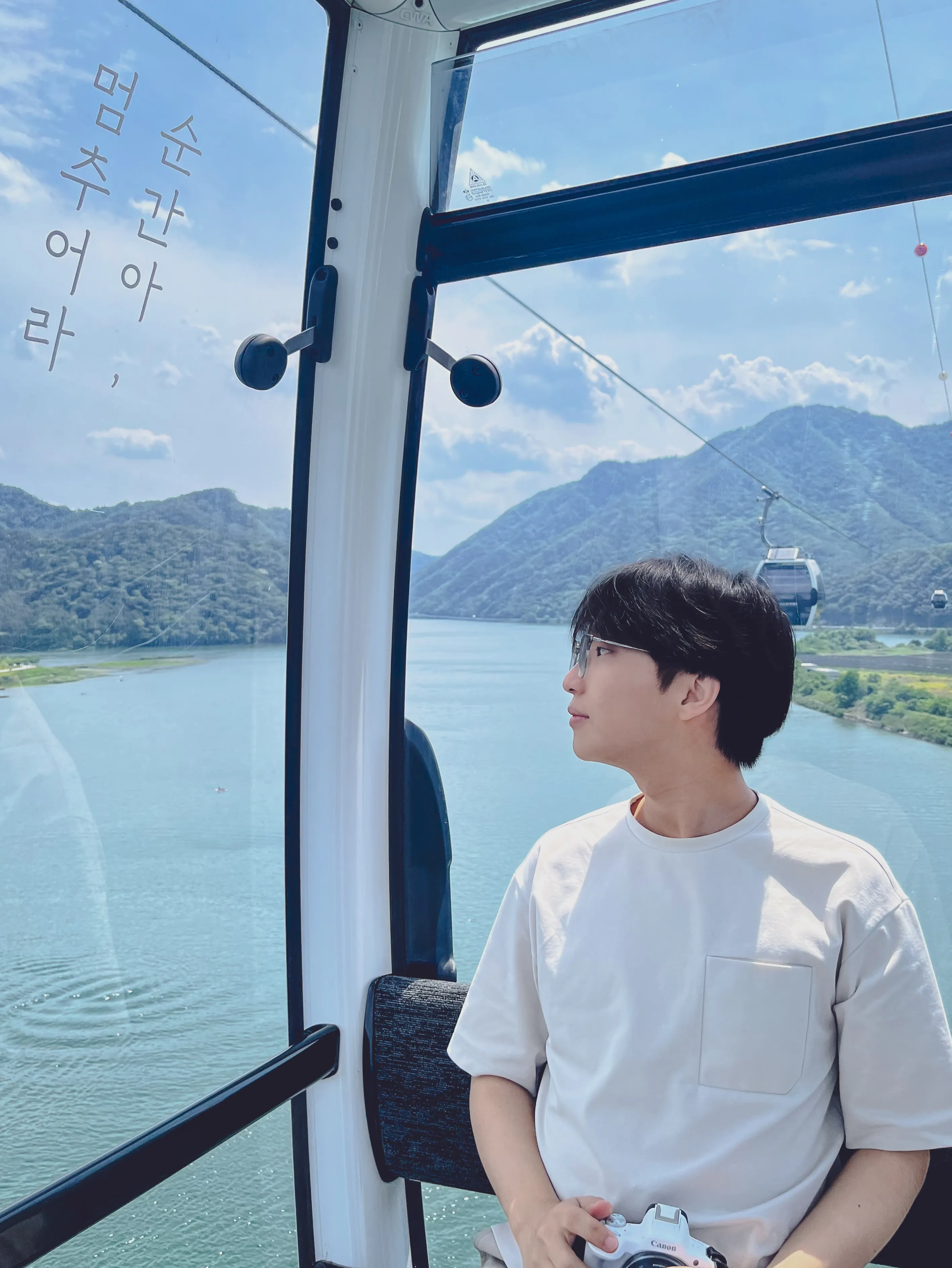 Me seated in a cable car, holding a camera, with a lake and mountains in the background