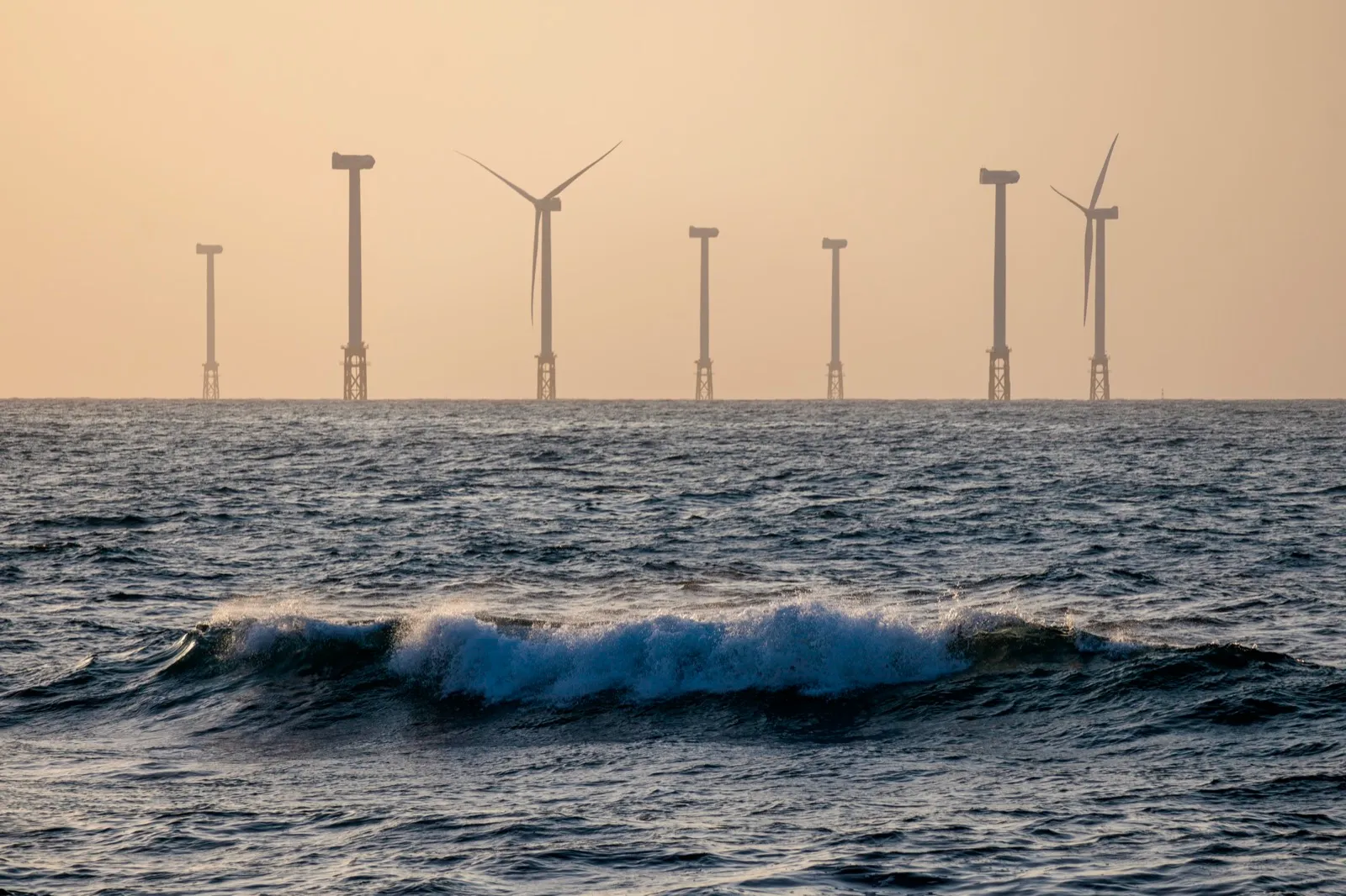 Offshore wind turbines lined up beyond dark ocean waves in hazy evening light