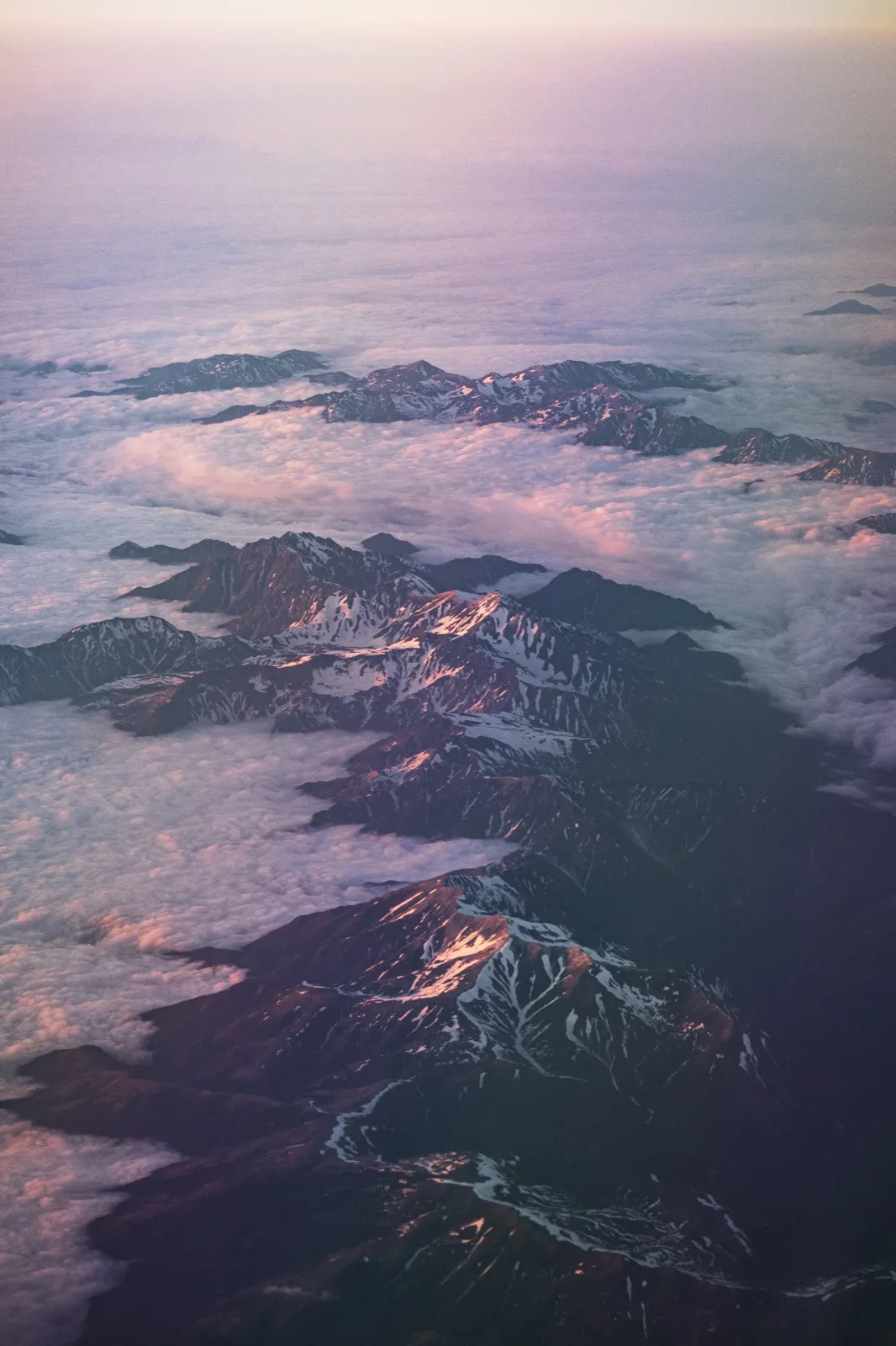 Snow-streaked mountain ridges seen from an airplane above a layer of pink evening clouds